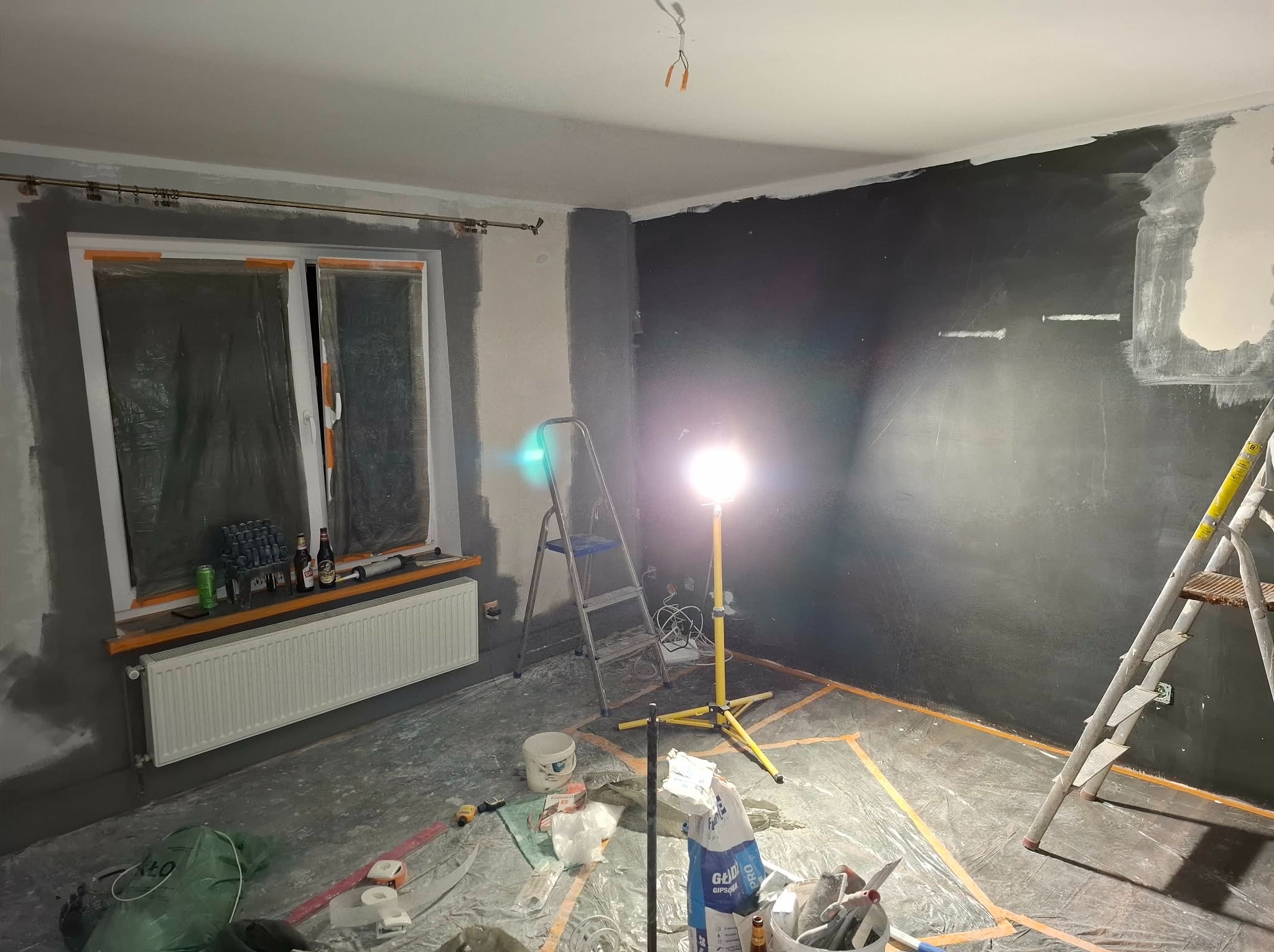 Pokój w trakcie malowania na ciemny kolor. Widoczne drabiny, folia ochronna, puszki z farbą i oświetlenie budowlane. Ściany częściowo pomalowane, okno zabezpieczone folią.