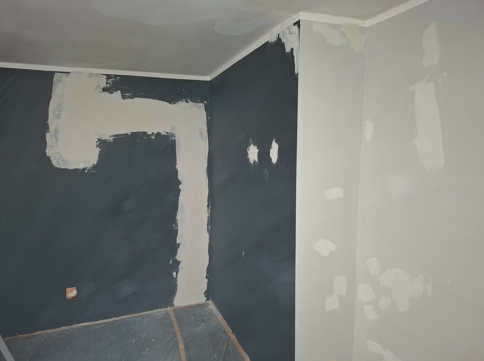 Pokój w trakcie szpachlowania: ciemna ściana z fragmentami nałożonej gładzi, jasna ściana z punktowymi poprawkami. Podłoga zabezpieczona folią.