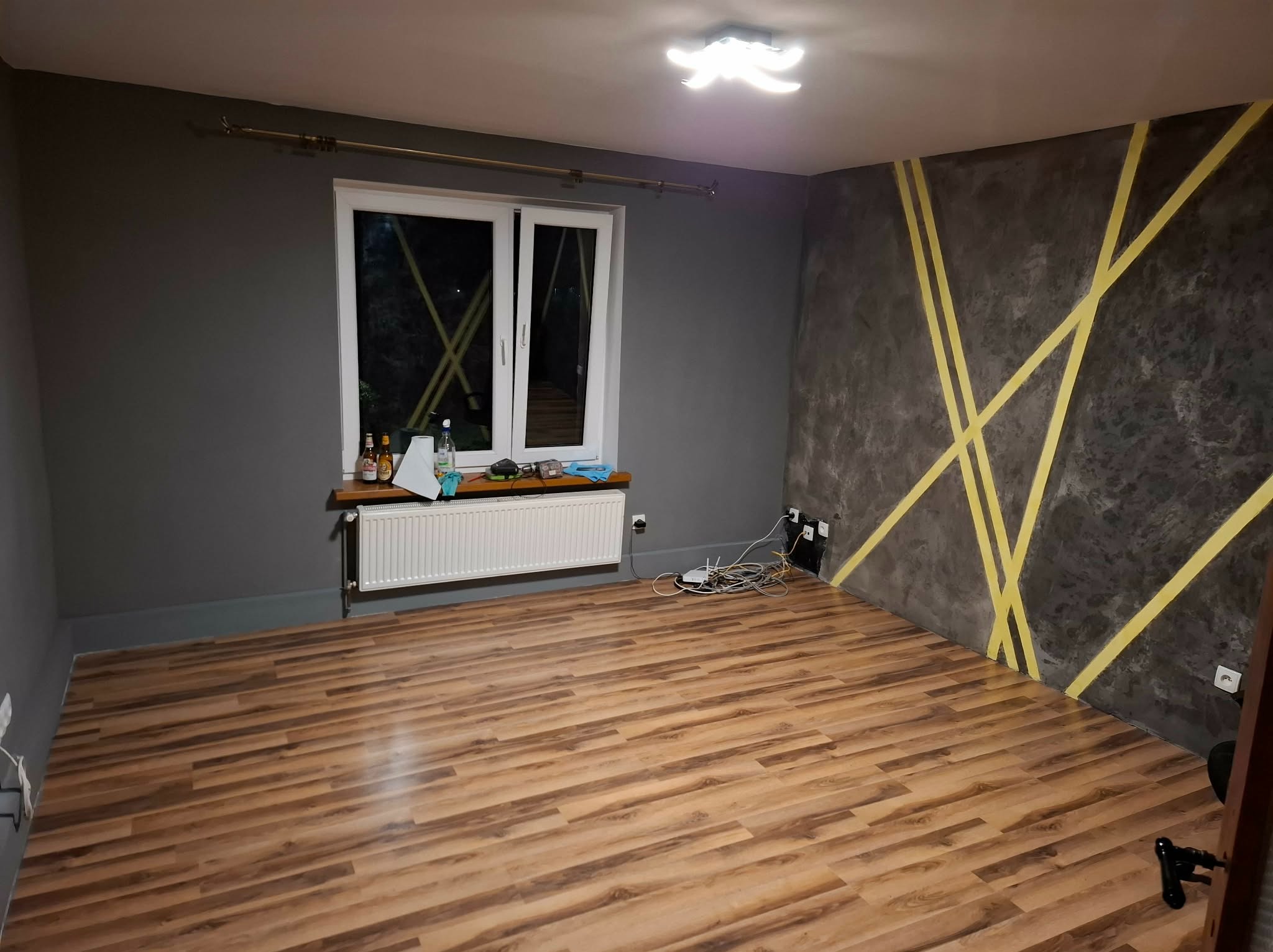 Szare wnętrze pokoju po malowaniu z dekoracyjnymi, żółtymi pasami na ścianie. Na podłodze panele, okno z grzejnikiem. Widoczne gniazdka i kable.