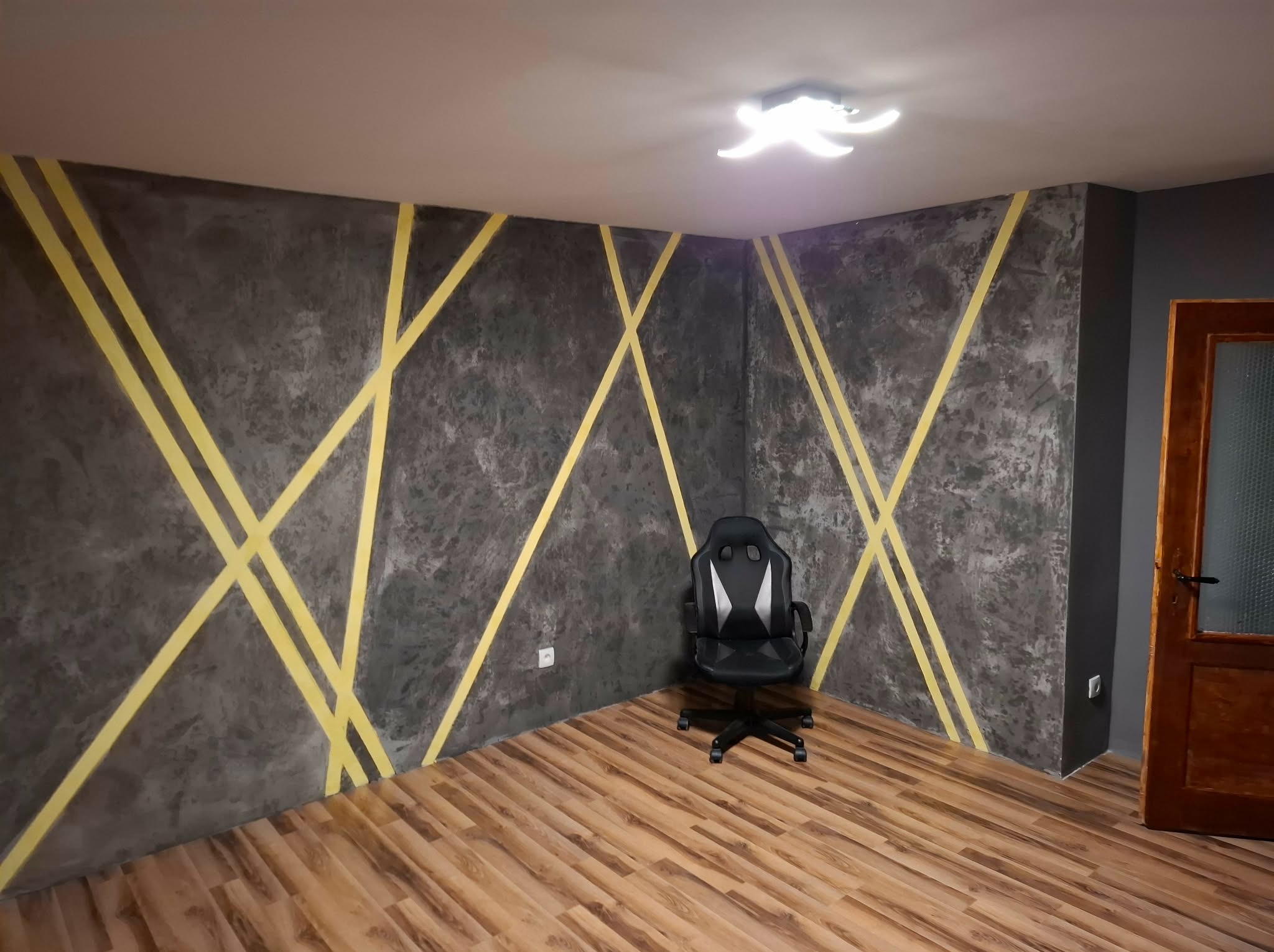 Pokój po malowaniu ścian z efektem betonu i żółtymi pasami. Na podłodze panele, w rogu krzesło gamingowe. Widoczne gniazdka elektryczne i oświetlenie sufitowe.