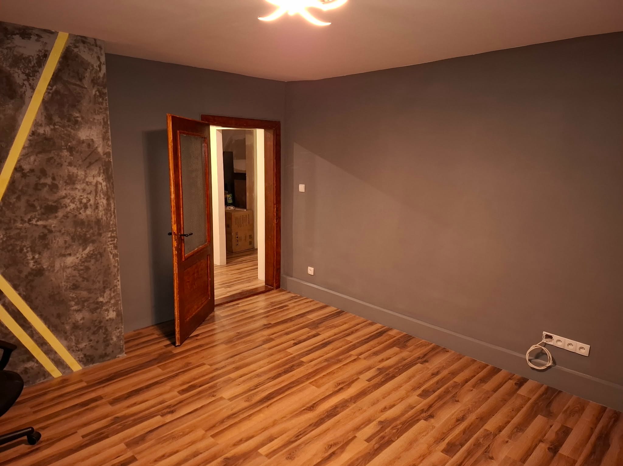 Pokój po remoncie: szare ściany, drewniana podłoga, otwarte drzwi z szybą. Na ścianie dekoracyjny beton z żółtymi pasami. Widoczne gniazdka elektryczne.