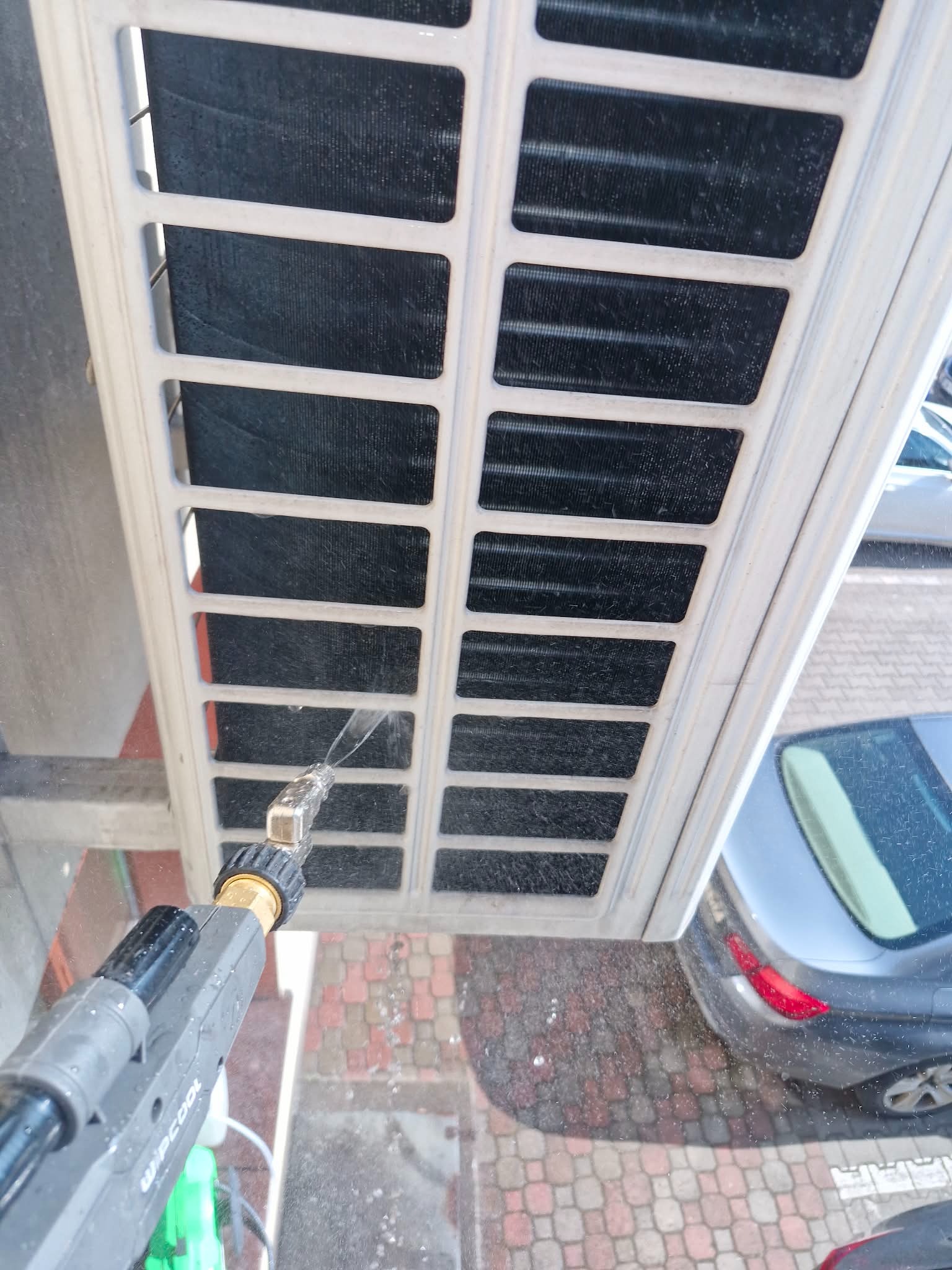 Czyszczenie klimatyzatora myjką ciśnieniową z bliska. Woda spryskuje filtry urządzenia na tle kostki brukowej i zaparkowanego samochodu.