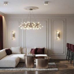 M2 Projekt - Elegancki salon z narożną sofą, stolikami kawowymi w kolorze złota i designerskim żyrandolem. Ściany zdobione sztukaterią, burgundowe akcenty w postaci zasłon i krzeseł barowych.