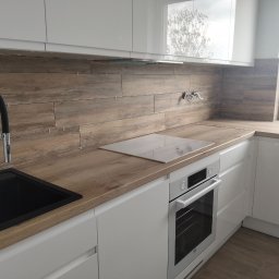 NAKMAR - Nowoczesna kuchnia z białymi szafkami i drewnopodobną okładziną ścienną w trakcie montażu, widoczne krzyżyki glazurnicze. Czarny zlew i chromowana bateria.