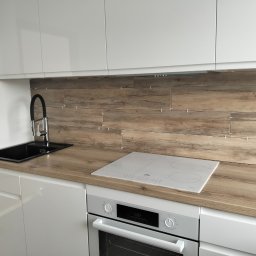 NAKMAR - Nowoczesna kuchnia z białymi szafkami i drewnopodobną okładziną ścienną w trakcie montażu, widoczne krzyżyki dystansowe. Czarny zlew i biała płyta indukcyjna.