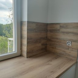 NAKMAR - Montaż płytek imitujących drewno w rogu pomieszczenia, widoczne krzyżyki dystansowe, gniazdko elektryczne i fragment okna z widokiem na zieleń.