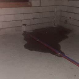 Surowy stan: Instalacja wodna w trakcie prac w pomieszczeniu z surowymi ścianami z betonu komórkowego. Widoczne rury i ślady po wycieku wody na posadzce.