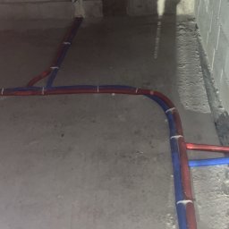 Instalacja rur PEX (czerwonych i niebieskich) na podłodze w surowym stanie, przymocowanych białymi uchwytami, z widocznymi bloczkami betonowymi w tle.