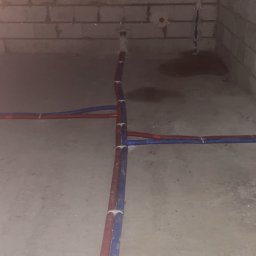 Surowy stan: Instalacja rur (czerwona i niebieska) na podłodze w pomieszczeniu z surowymi ścianami z pustaków. Widoczne punkty przyłączeniowe na ścianie.