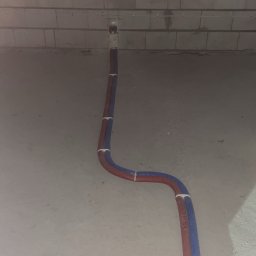 Dwie rury (czerwona i niebieska) prowadzone po podłodze w surowym wnętrzu, przymocowane do ściany z pustaków. Widoczne mocowania rur.