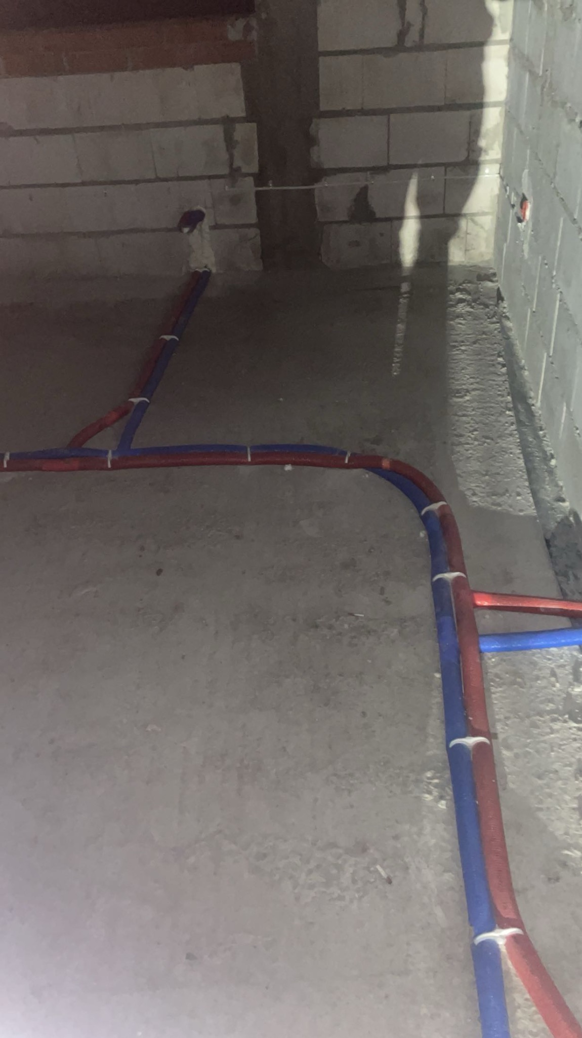 Instalacja rur PEX (czerwonych i niebieskich) na podłodze w surowym stanie, przymocowanych białymi uchwytami, z widocznymi bloczkami betonowymi w tle.