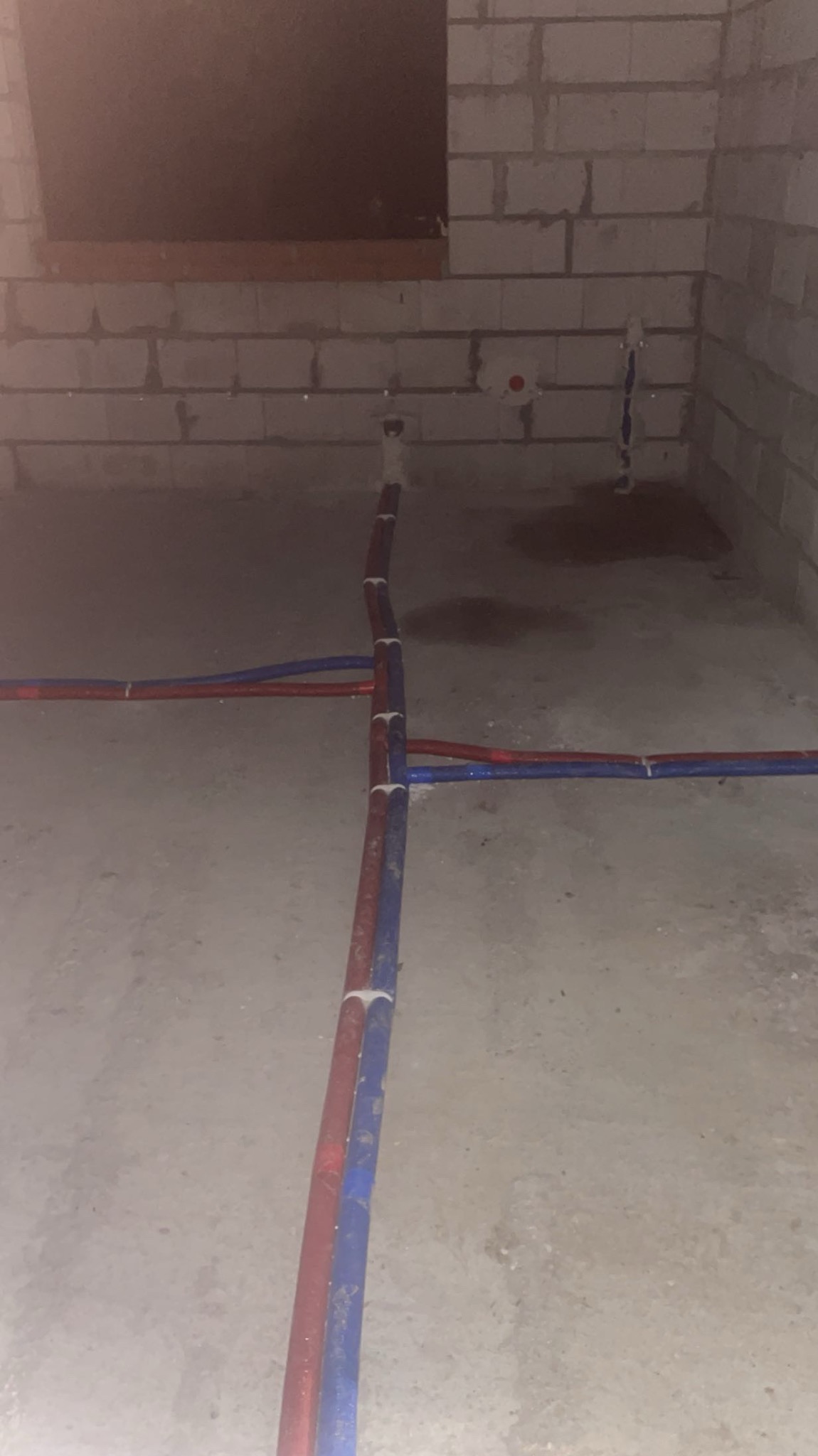 Surowy stan: Instalacja rur (czerwona i niebieska) na podłodze w pomieszczeniu z surowymi ścianami z pustaków. Widoczne punkty przyłączeniowe na ścianie.