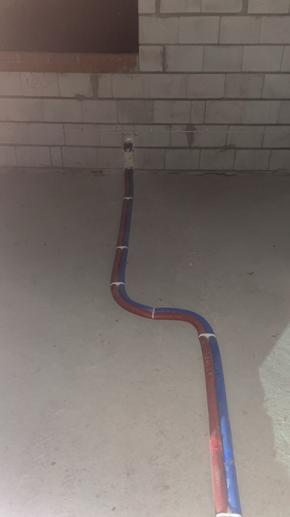 Dwie rury (czerwona i niebieska) prowadzone po podłodze w surowym wnętrzu, przymocowane do ściany z pustaków. Widoczne mocowania rur.