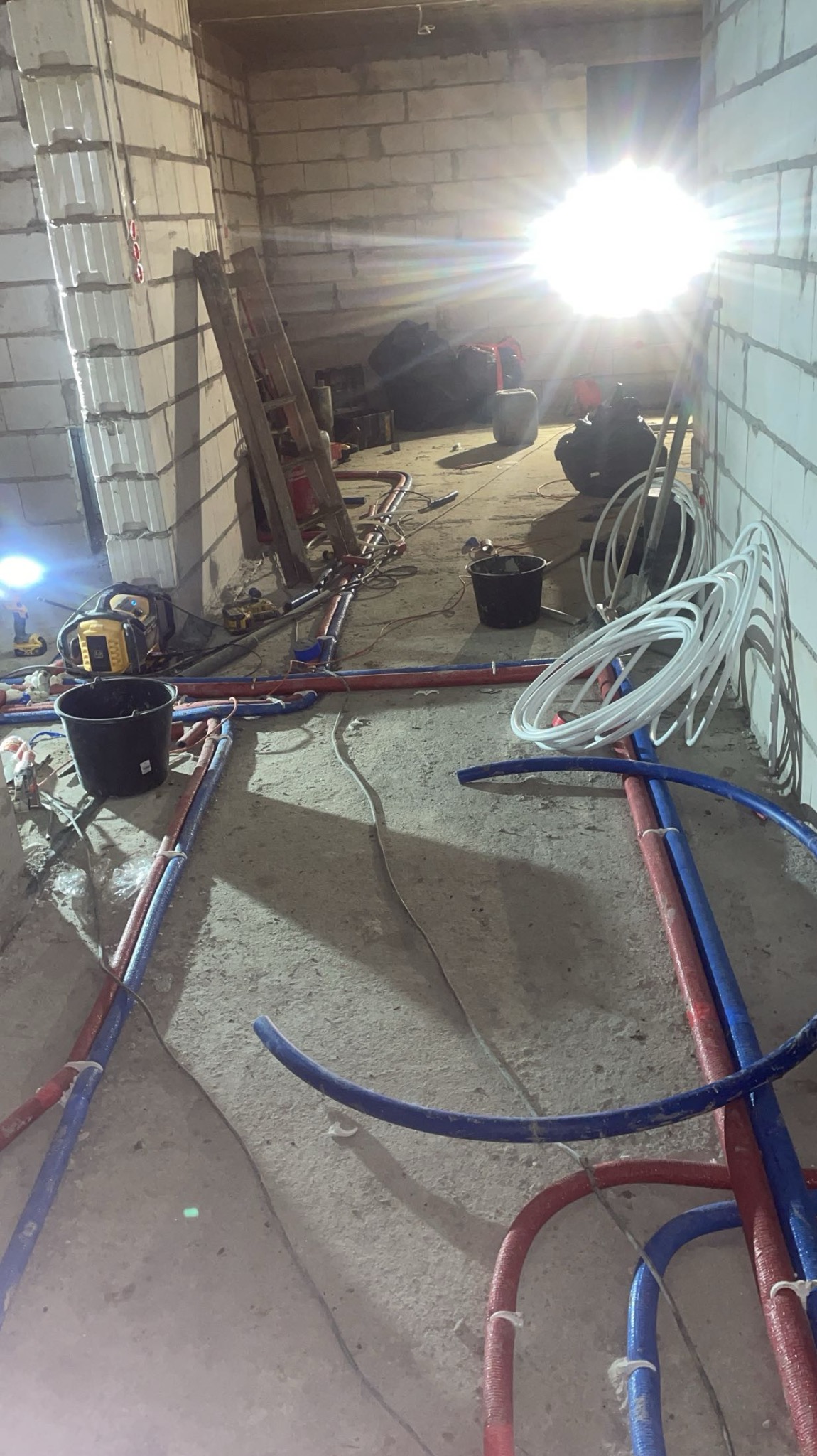Instalacja rur PEX w surowym wnętrzu. Widoczne narzędzia, drabina i wiadro. Rury czerwone i niebieskie układane na podłodze, białe zwinięte w kręgi. Ostre światło w tle.
