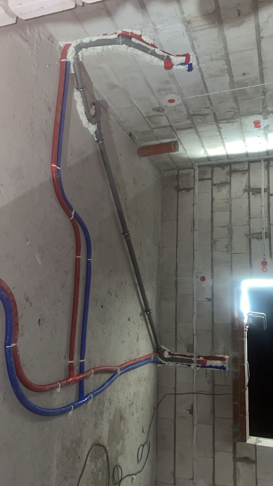 Surowe wnętrze z instalacją hydrauliczną: rury czerwone i niebieskie prowadzone po ścianie i suficie, zabezpieczone pianką montażową. Widoczne przewody elektryczne.