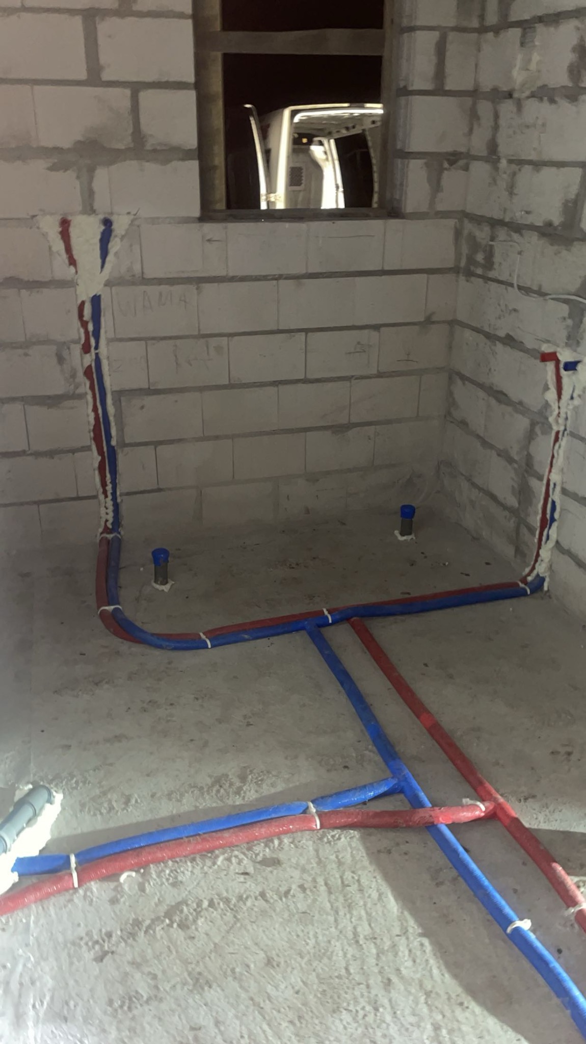Instalacja wodna z rurami czerwonymi i niebieskimi, zabezpieczonymi pianką montażową przy ścianach z betonu komórkowego. Widoczne piony i poziomy układ rur.