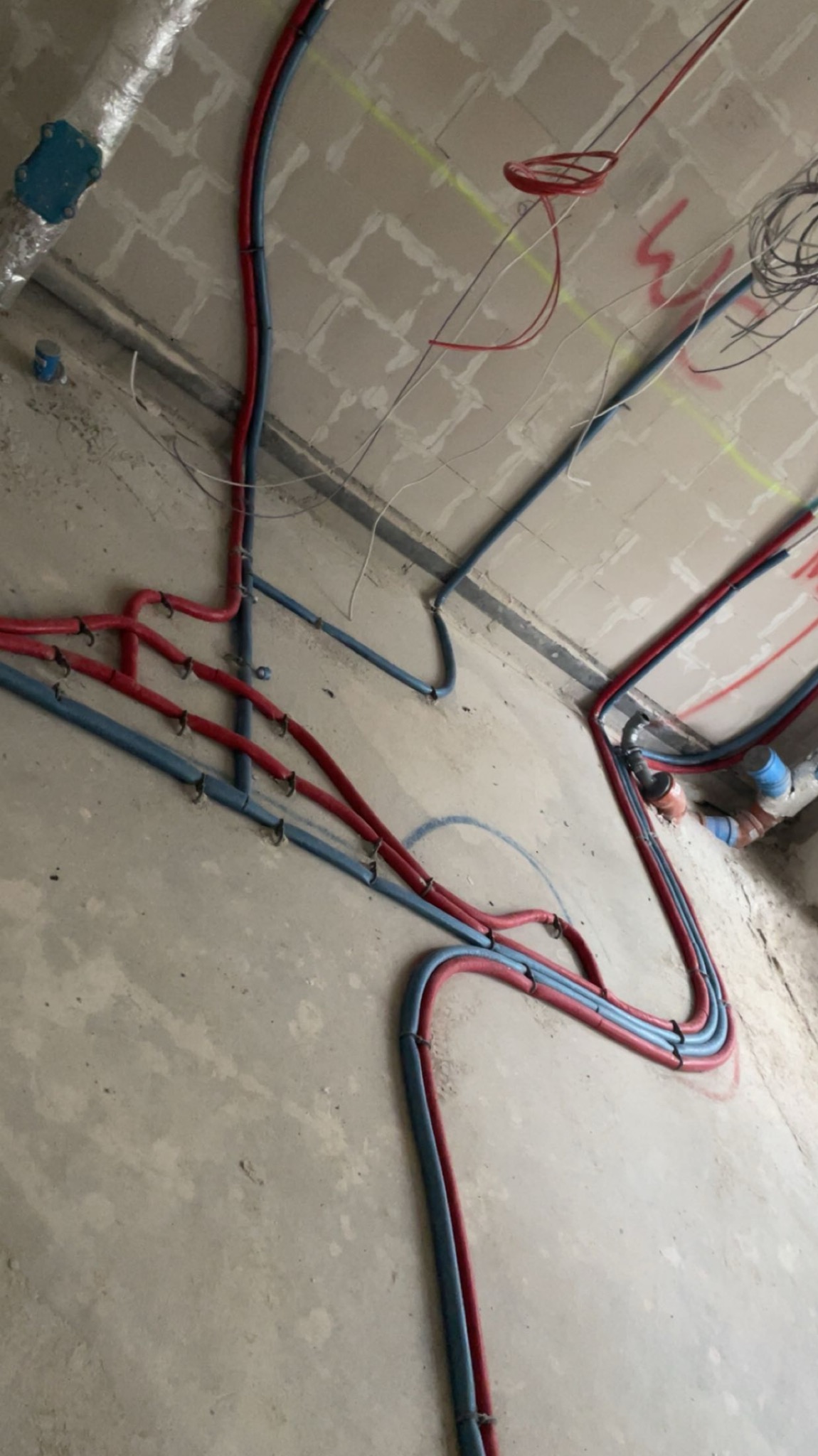 Wnętrze w trakcie prac: czerwone i niebieskie rury instalacji biegnące po podłodze i suficie, mocowane do ściany. Widoczne ślady po instalacjach elektrycznych.
