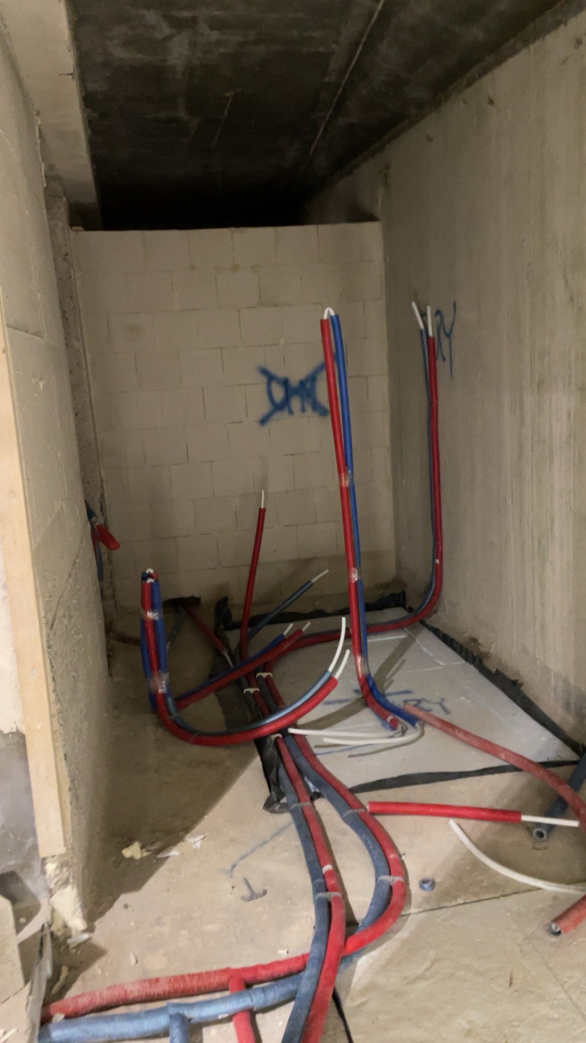 Instalacja ogrzewania podłogowego w surowym wnętrzu. Czerwono-niebieskie rury ułożone na izolacji, widoczne ściany z betonu komórkowego i surowy strop.