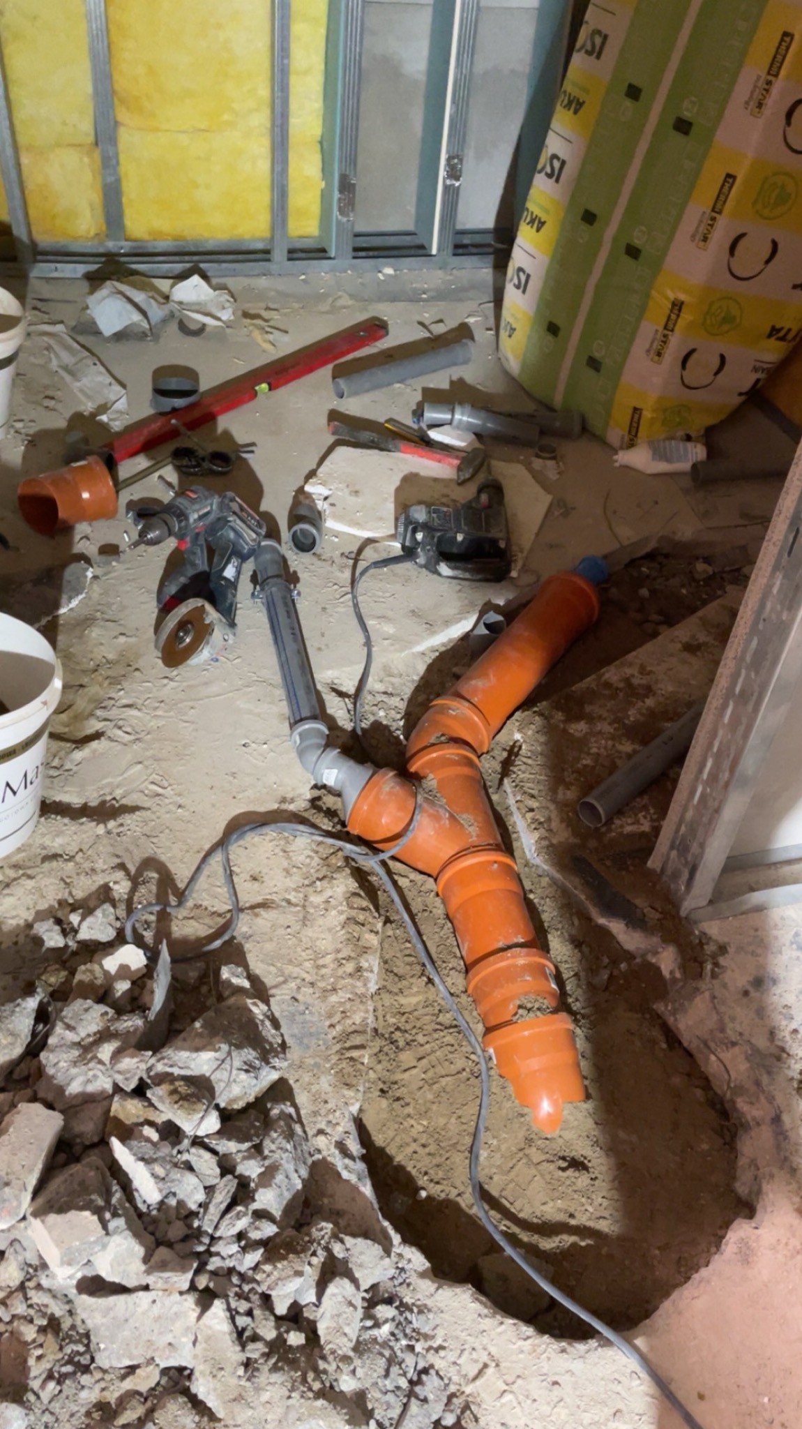 Pomarańczowe rury kanalizacyjne w wykopie, narzędzia hydrauliczne i materiały budowlane w tle. Widok z góry na plac budowy w trakcie prac instalacyjnych.