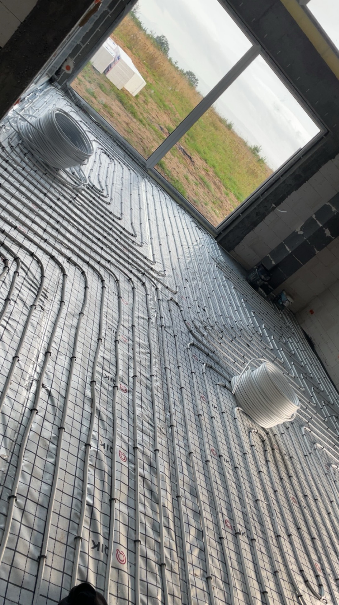 Instalacja ogrzewania podłogowego na siatce, widok z góry, z rolkami rur i oknem dachowym w tle. Etap przed wylaniem posadzki, widoczne detale mocowań rur.