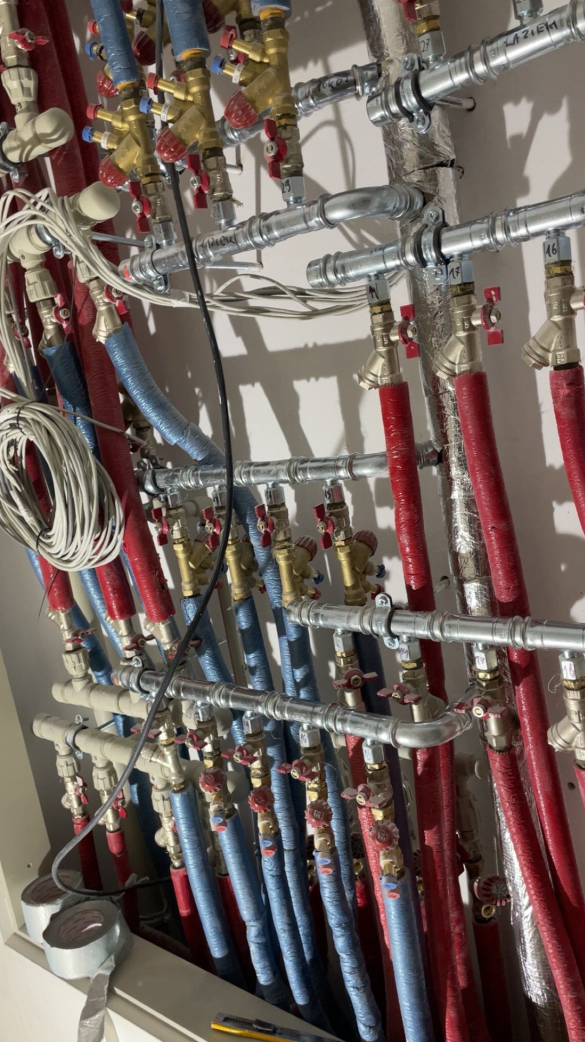Rozbudowana instalacja hydrauliczna z rurami w izolacji (czerwona i niebieska), zaworami i przewodami. Widoczne oznaczenia na rurach.