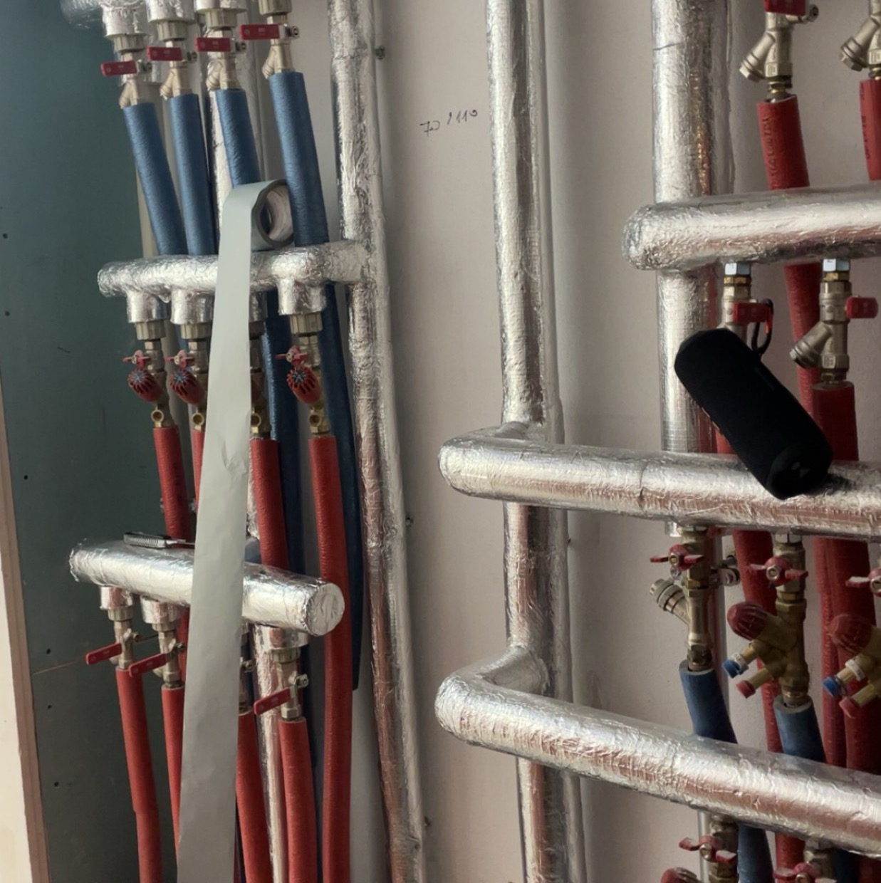 Izolowane rury instalacji grzewczej z zaworami, zabezpieczone folią aluminiową, prowadzone wzdłuż ściany. Widoczne rury w kolorze czerwonym i niebieskim.