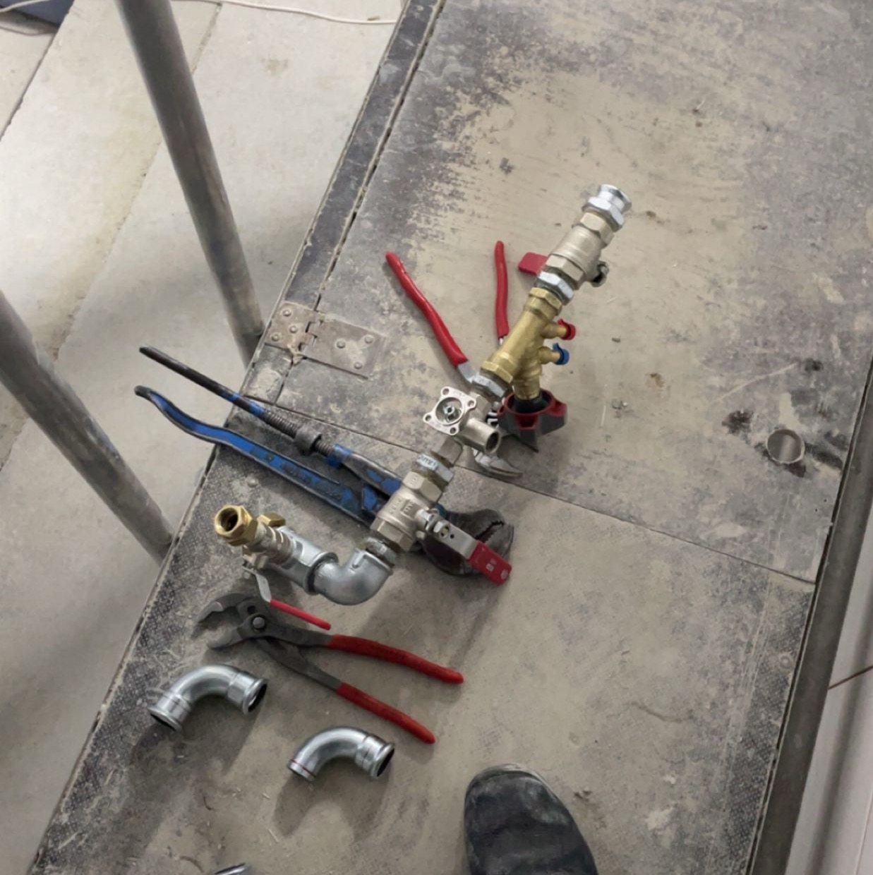 Przygotowane elementy instalacji hydraulicznej z narzędziami na brudnej powierzchni roboczej. Widoczne złączki, rury, klucze nastawne i fragment buta.