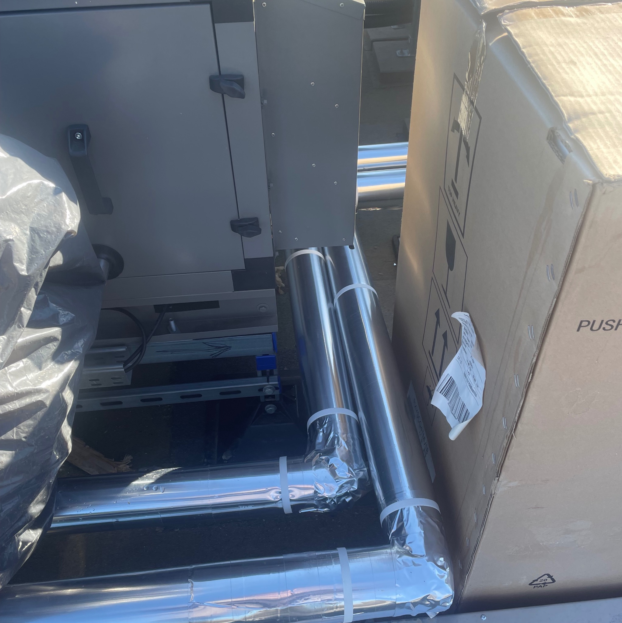 Fragment instalacji wentylacyjnej z izolowanymi rurami, widoczne elementy centrali wentylacyjnej i kartonowe opakowanie. Ujęcie z bliska, z góry.