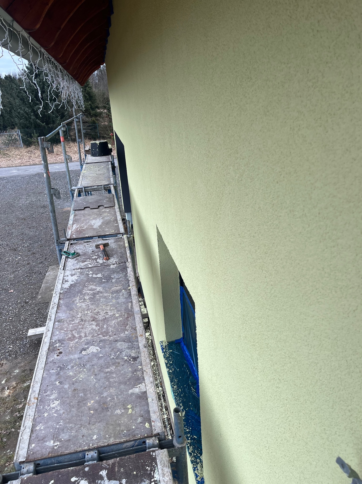 Ściana budynku po nałożeniu strukturalnego tynku w odcieniach beżu, widoczna z rusztowania. Okno zabezpieczone folią. W tle drzewa i fragment drogi.