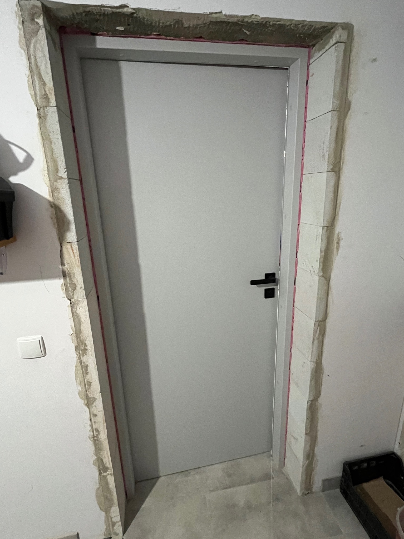 Białe drzwi wewnętrzne z czarną klamką, osadzone w surowym otworze z widoczną pianką montażową i resztkami zaprawy, na tle jasnej ściany i podłogi.