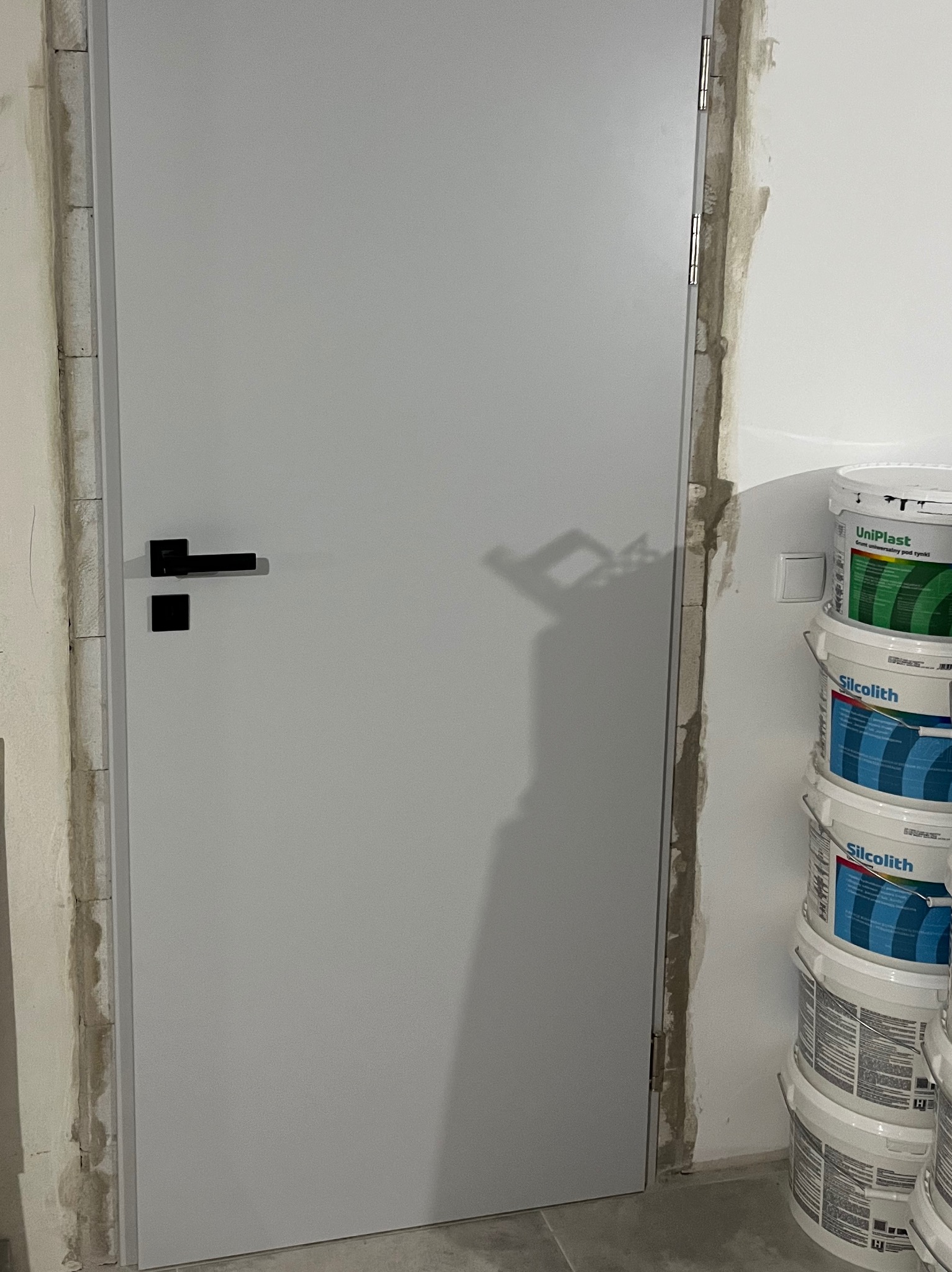 Zamknięte, białe drzwi z czarną klamką w surowym otoczeniu. Widoczne ślady po pracach wykończeniowych i wiaderka z farbą. Minimalistyczny design.
