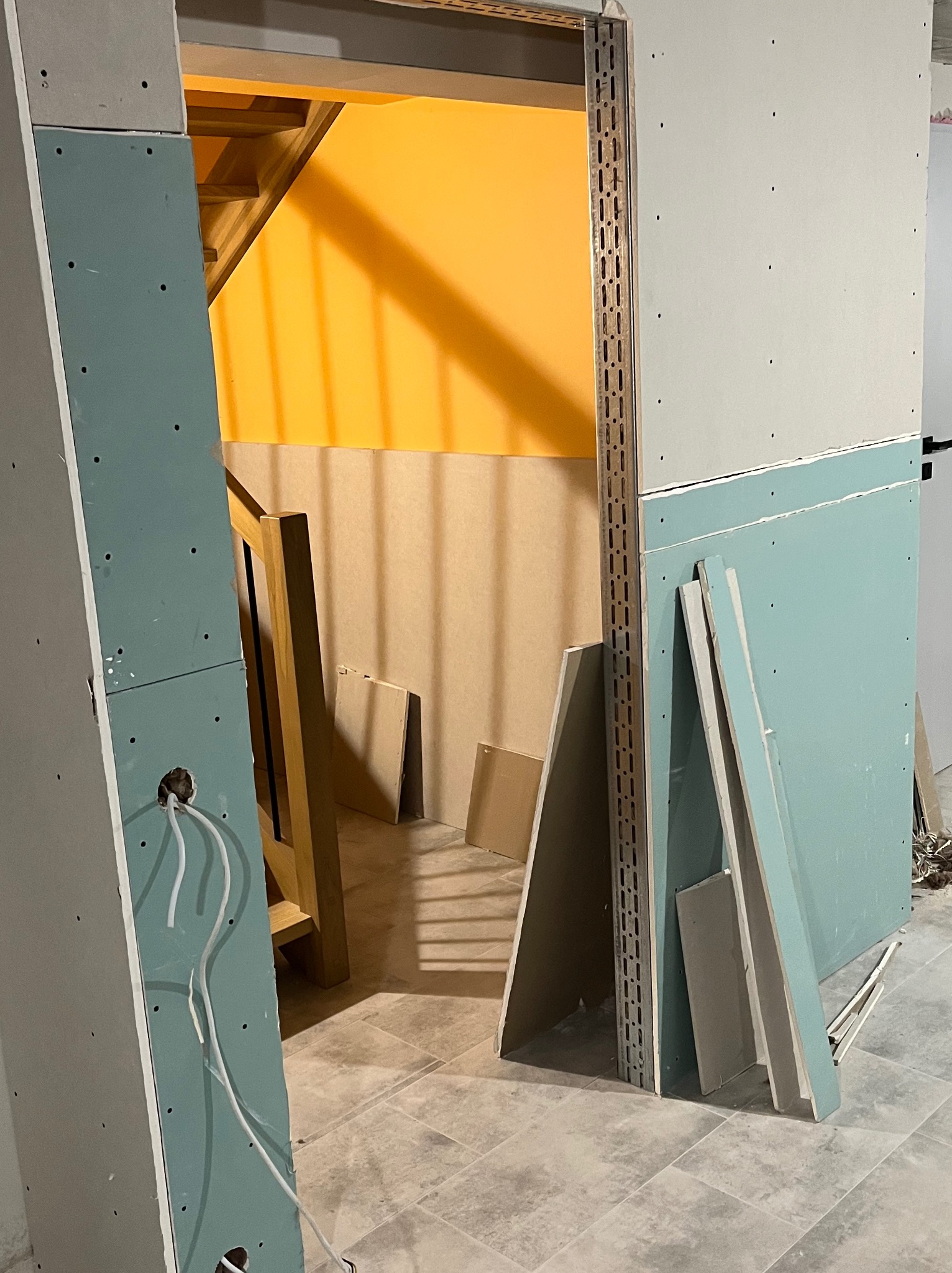 Montaż płyt karton-gips na metalowym stelażu, widoczne przewody elektryczne i fragment schodów z pomarańczową ścianą w tle. Prace wykończeniowe w surowym wnętrzu.