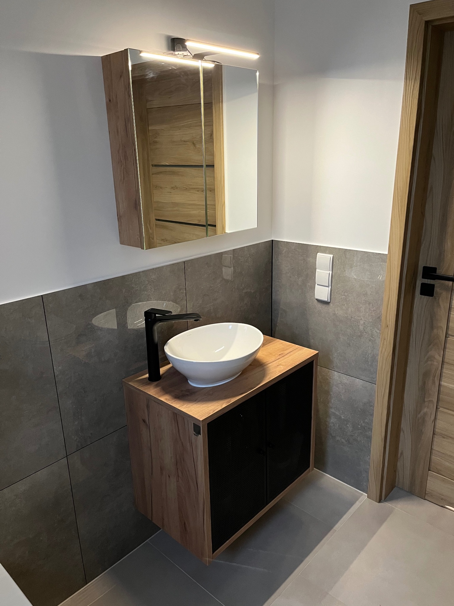 Nowoczesna łazienka w Ochodzach: szare płytki, drewniane szafki z umywalką nablatową i lustro z oświetleniem. Minimalistyczny design i funkcjonalność.