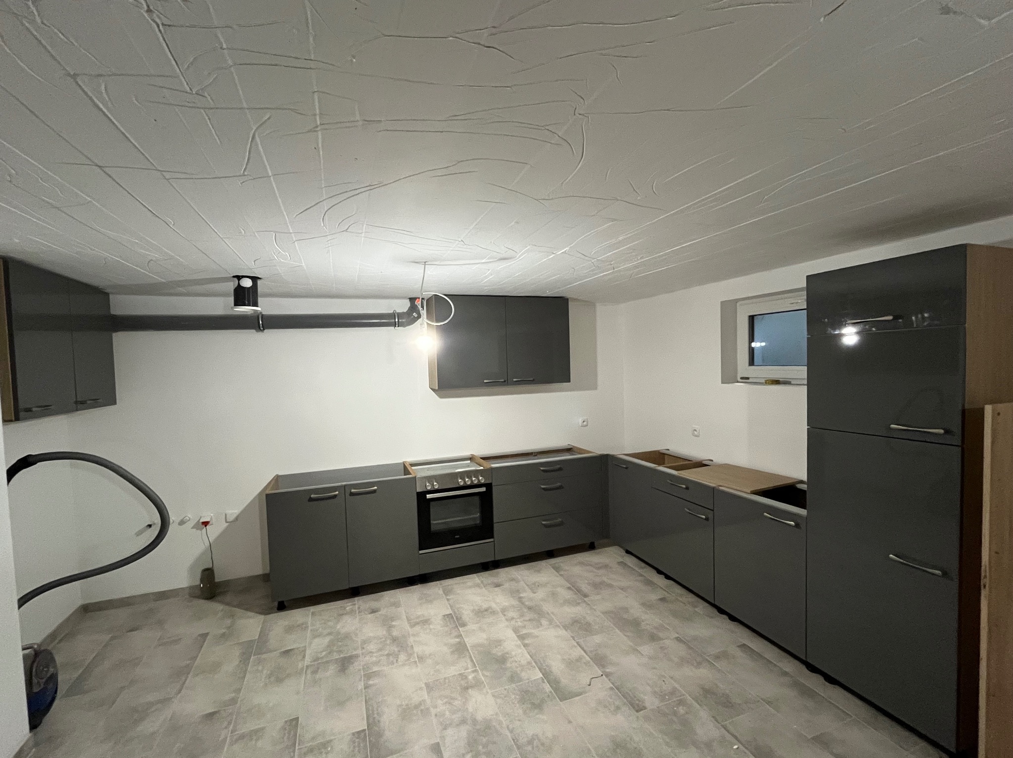 Szara, nowoczesna zabudowa kuchenna w trakcie montażu, widoczny odkurzacz, surowy sufit i płytki podłogowe. Brak blatów i wykończeń.