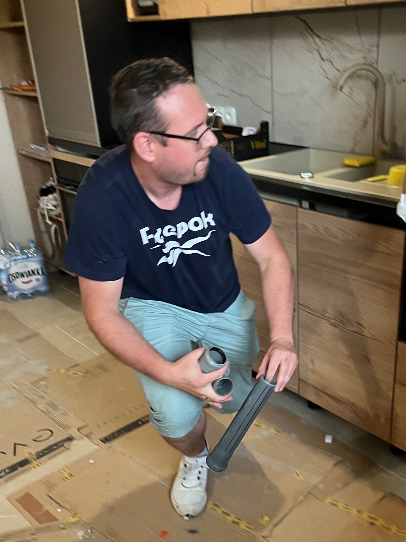 Mężczyzna w okularach klęczy w kuchni, trzymając rury kanalizacyjne. Widoczne szafki, zlew i kartony na podłodze. Prace instalacyjne w trakcie realizacji.