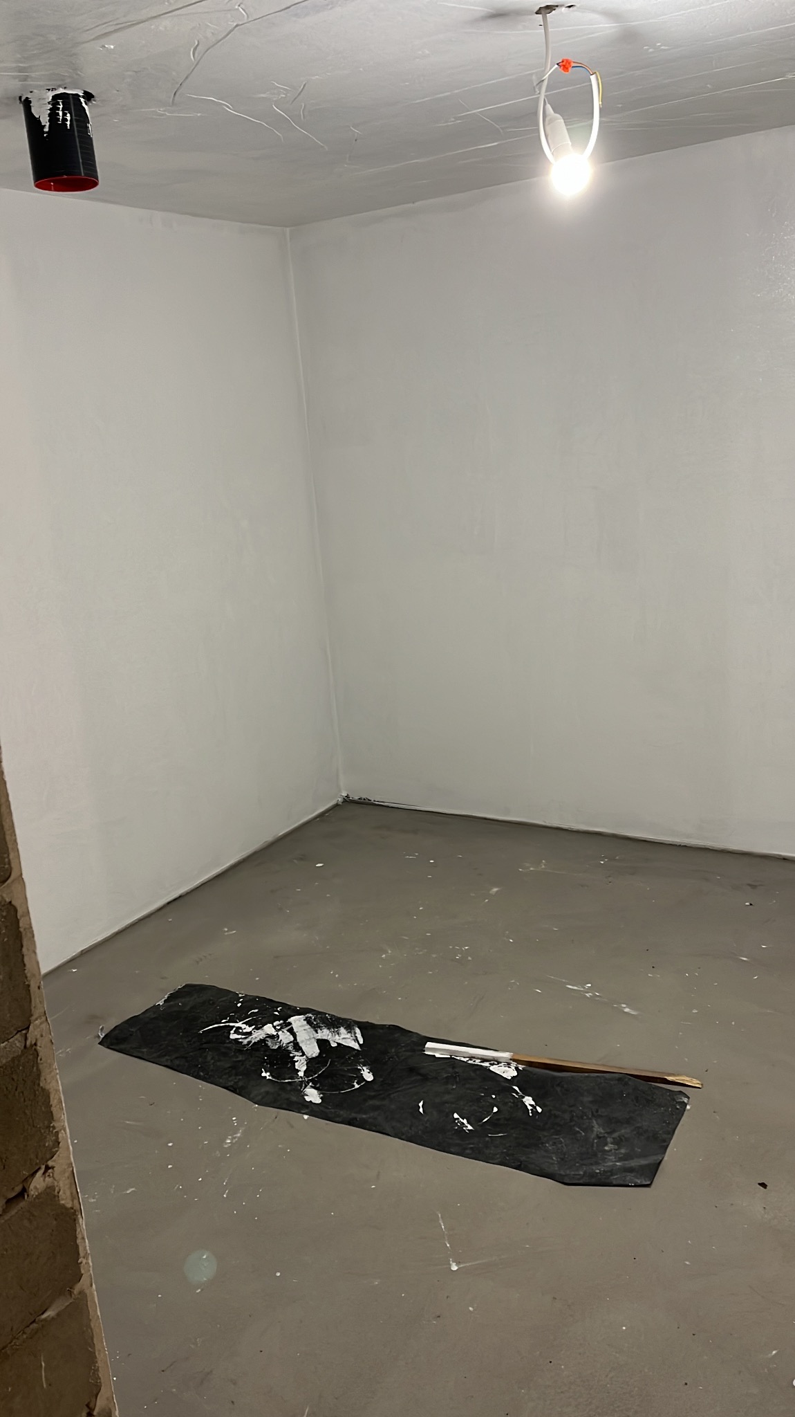 Wnętrze po szpachlowaniu: gładkie, białe ściany i sufit, zabezpieczona podłoga z widocznymi śladami farby. Przy suficie widoczna instalacja elektryczna.