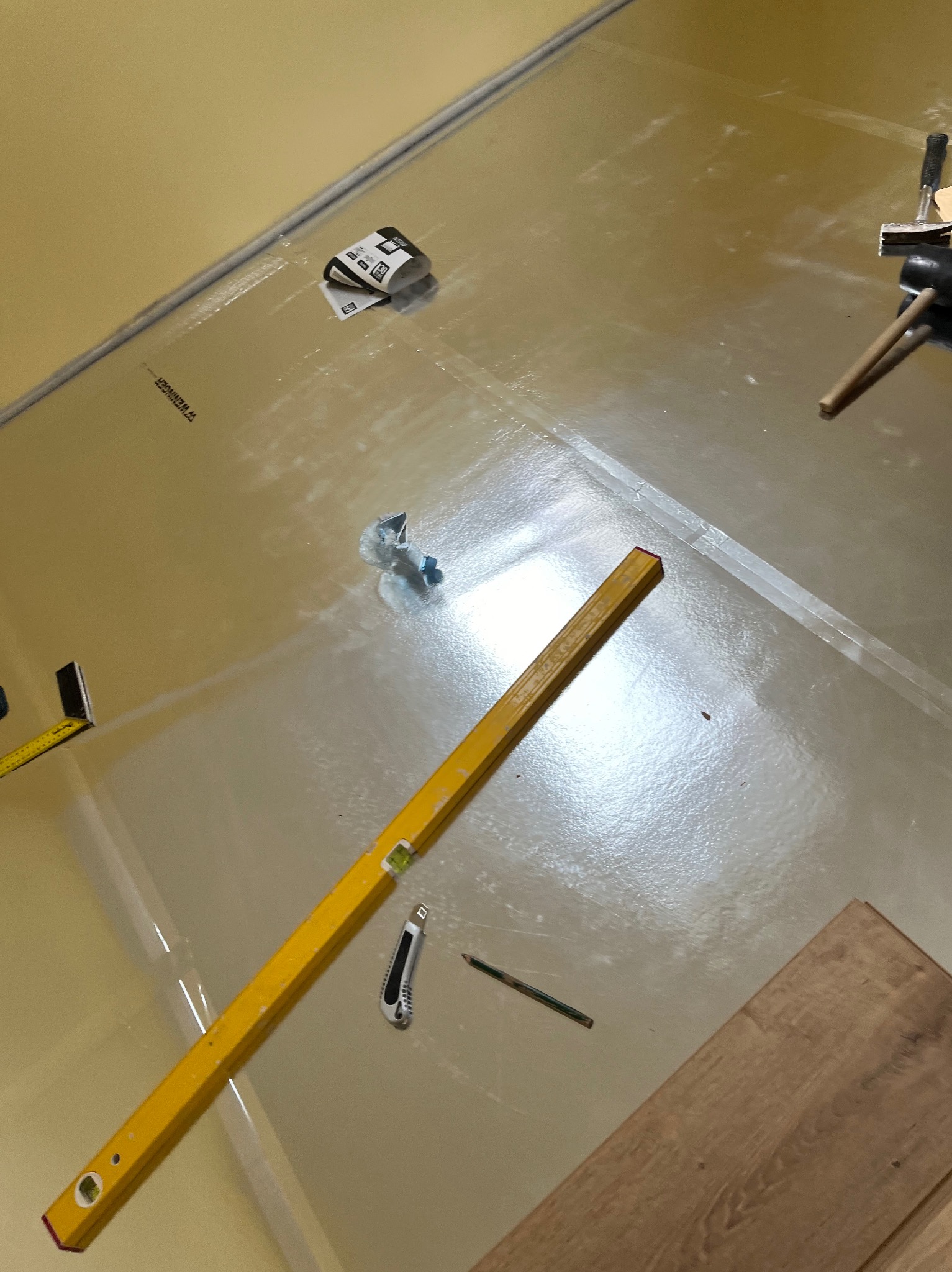 Przygotowanie podłoża pod parkiet: folia, poziomica, nóż, ołówek. Widoczne narzędzia i materiały na błyszczącej folii izolacyjnej, gotowe do układania paneli.