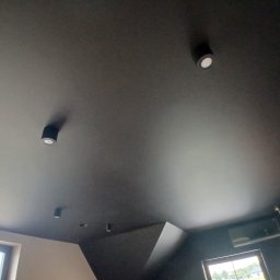 Investbud - Czarny sufit z punktowym oświetleniem LED, widok z dołu. Minimalistyczny design wnętrza z oknami w tle. Nietypowy kąt ujęcia podkreśla gładką powierzchnię sufitu.