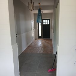 Investbud - Korytarz po remoncie: szara podłoga z płytek, ceglana posadzka w jodełkę, białe panele ścienne. W tle czarne drzwi wejściowe i wisząca lampa w folii ochronnej.