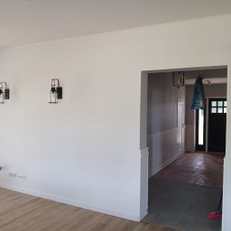 Investbud - Białe, poziome panele ścienne z dwoma czarnymi kinkietami. Widoczne instalacje elektryczne, podłoga z paneli i przejście do innego pomieszczenia z ceglaną podłogą.