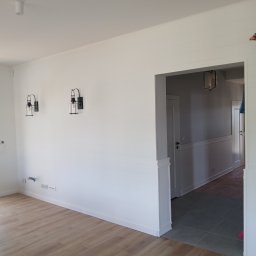 Investbud - Białe, poziome panele ścienne z czarnymi kinkietami w wykończonym wnętrzu. Podłoga z paneli imitujących drewno, widok na korytarz z szarą podłogą.