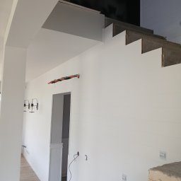 Investbud - Białe wnętrze w trakcie wykończenia: ściany z boazerią, surowe schody betonowe, lampy ścienne i widoczne instalacje elektryczne. Wnętrze w stylu nowoczesnym.