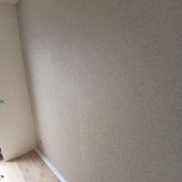 Investbud - Beżowa ściana po szpachlowaniu i malowaniu, widoczna faktura. Podłoga drewniana z listwami przypodłogowymi. Gniazdko elektryczne na ścianie. Ujęcie z dołu, ukośne.