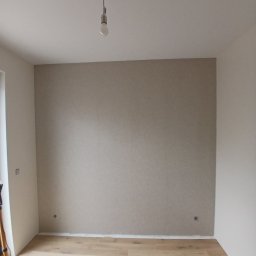 Investbud - Pokój po szpachlowaniu i malowaniu jednej ściany na beżowo. Widoczna podłoga z paneli, surowa żarówka i fragment statywu. Ściany w odcieniach bieli i beżu.