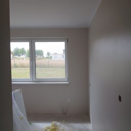 Investbud - Świeżo pomalowane wnętrze pokoju z oknem, widoczne zabezpieczenia podłogi i ściany. Gładkie, jasne ściany i sufit. Przygotowanie do dalszych prac wykończeniowych.