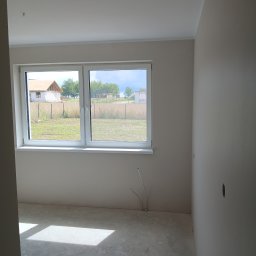 Investbud - Jasne wnętrze po malowaniu ścian na beżowo, z widocznym oknem i surową podłogą. W tle krajobraz z domem w budowie. Widoczne instalacje elektryczne przy podłodze.