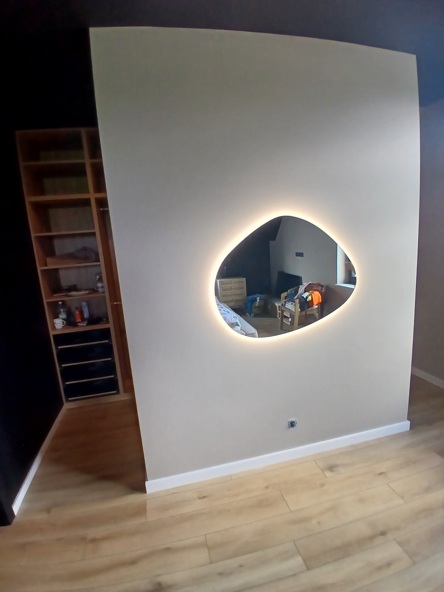 Ściana z asymetrycznym lustrem podświetlanym LED, odbijającym wnętrze pokoju. Parkiet na podłodze, widoczna szafa wnękowa. Minimalistyczny design.