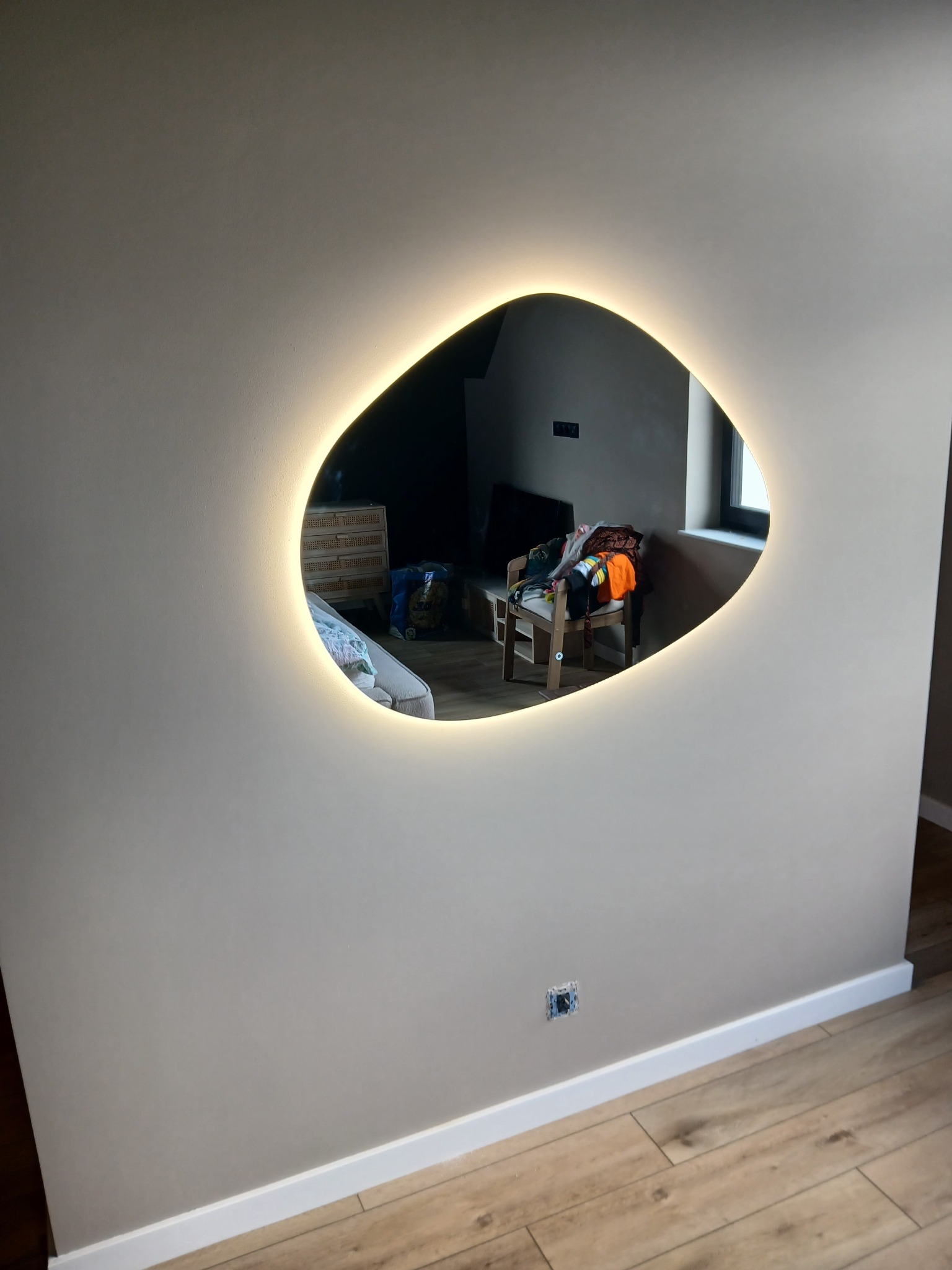 Narożnik pokoju z beżową ścianą, na której wisi asymetryczne lustro z podświetleniem LED. Widoczna podłoga z paneli i biała listwa przypodłogowa.