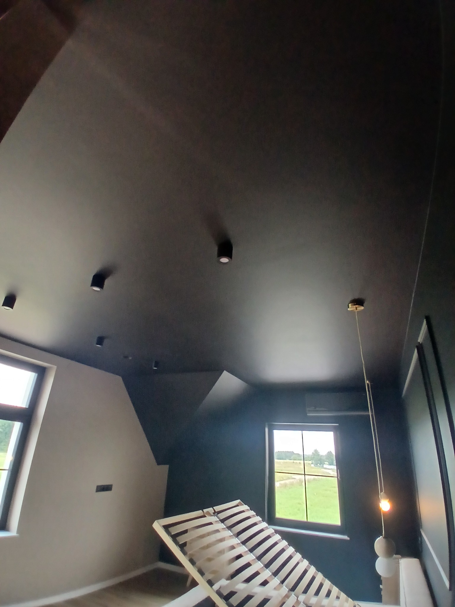 Czarne wykończenie sufitu w pokoju na poddaszu z oknami, widoczne oświetlenie punktowe i wisząca lampa. Częściowo widoczny stelaż łóżka.