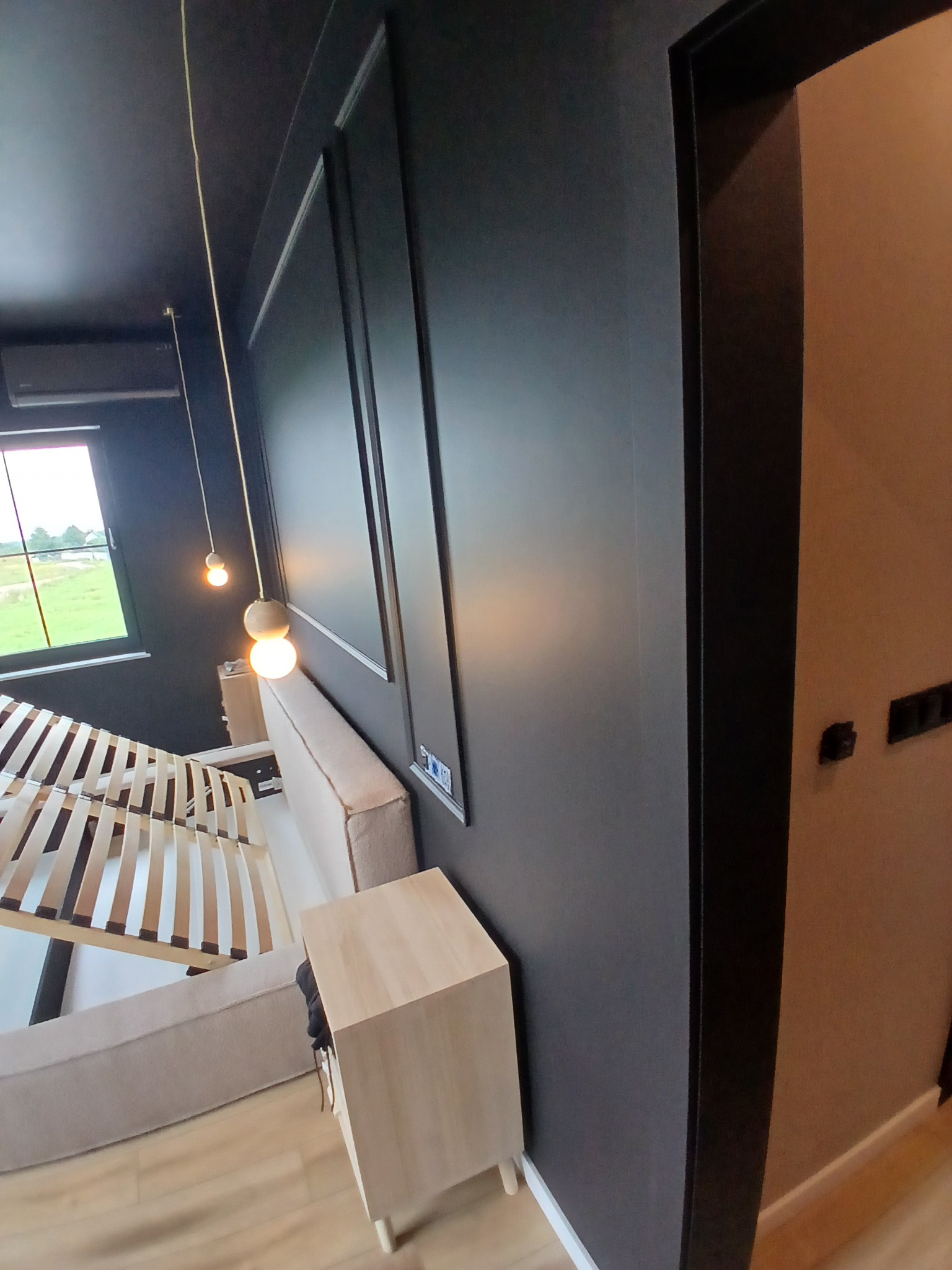 Nowoczesna sypialnia z czarnymi ścianami, listwami dekoracyjnymi i drewnianym łóżkiem. Widoczne lampy wiszące i szafka nocna. Minimalistyczny design wnętrza.